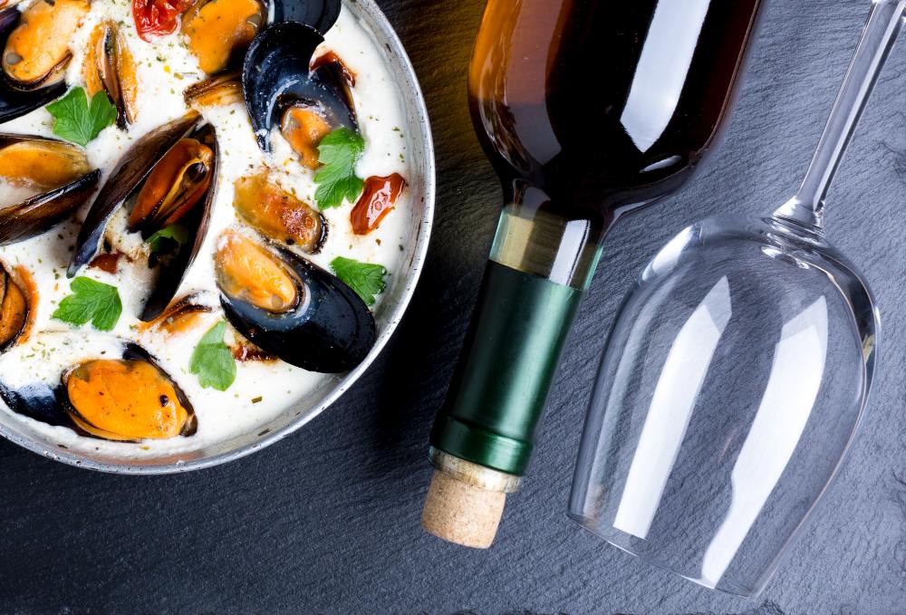 Assiette de moules à la crème avec tomates et persil frais, accompagnée d’une bouteille de vin blanc et d’un verre vide sur ardoise noire.