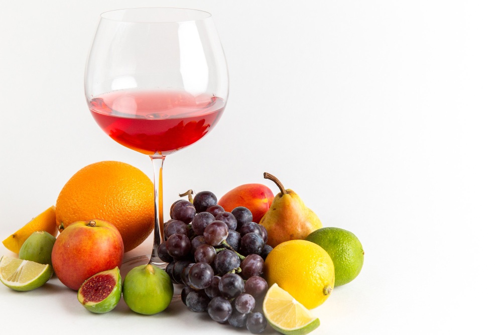 Verre de vin rouge entouré de fruits frais variés : orange, raisin, citron, figue, poire, pomme, lime et mangue sur fond blanc.