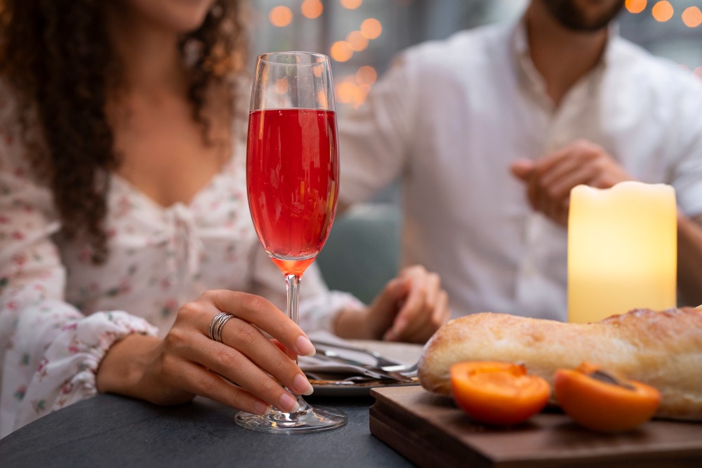 Femme tenant un verre de cocktail rouge dans un cadre romantique, avec une bougie allumée, du pain et des abricots posés sur la table.