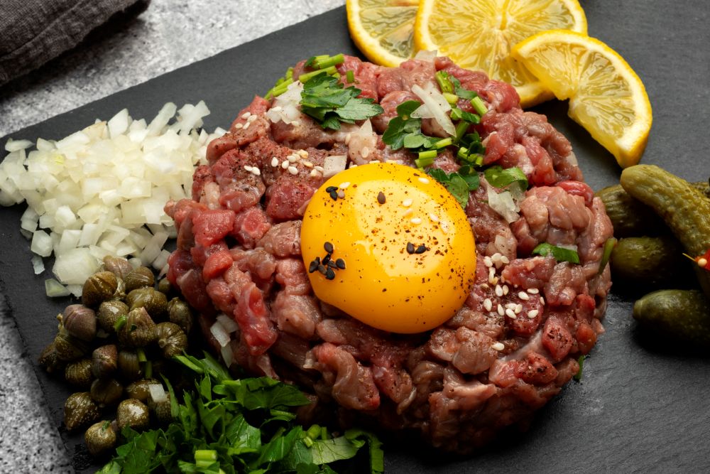 Steak tartare garnit d'un œuf, de câpres, cornichons, citrons, oignons, salé et poivré accompagné de quelques herbes.